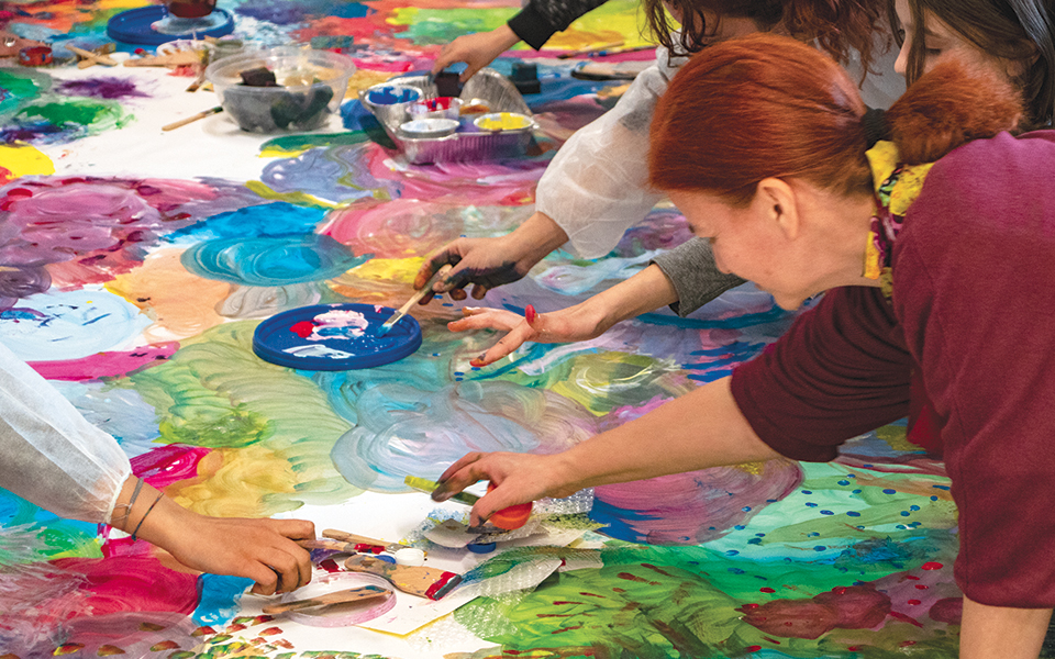 Artista dai capelli rossi seduta sul pavimento davanti a un dipinto insieme a molte persone colorato realizzato all'evento family art