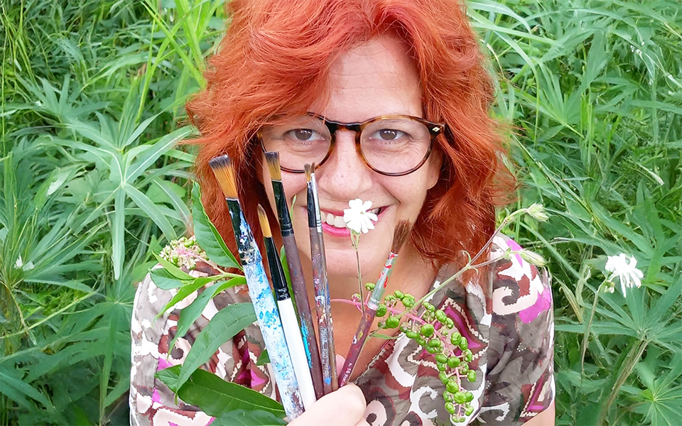 Artista dai capelli rossi con occhiali, sorridente, che tiene pennelli, fiori e piante in primo piano